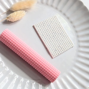 Puede incluir: Un objeto cilíndrico rosa texturizado y un objeto rectangular blanco con un patrón moteado descansan sobre un plato blanco acanalado. Dos tallos secos de hierba cola de conejo beige también están en el plato. Los objetos están dispuestos para una estética minimalista.