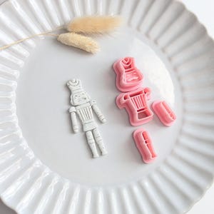 Könnte beinhalten: Eine weiße Nussknacker-Figur mit grauen Sprenkeln neben drei rosa Kunststoff-Ausstechformen. Die Formen sind als Kopf, Rumpf und Beine eines Nussknackers gestaltet, um die abgebildete zusammengesetzte Figur zu formen.