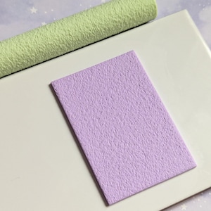 Puede incluir: Dos piezas de piel sintética texturizada en verde claro y lavanda. La pieza lavanda es rectangular y la pieza verde está enrollada.