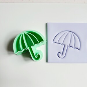 Puede incluir: Un cortador de galletas de plástico verde con forma de paraguas, junto a un sello de plástico morado con el mismo diseño.