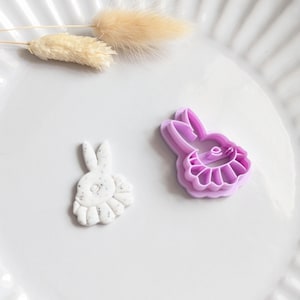 Puede incluir: Un plato de cerámica blanco contiene una pequeña figura de conejo moteado y un cortador de galletas con forma de conejo morado. Hierba beige seca y esponjosa acentúa la escena. Los artículos son probablemente para hornear con temática de Pascua o primavera.