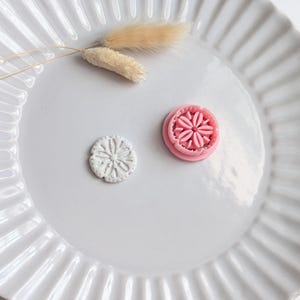 May include: A white ceramic plate with a fluted rim holds a small, white, flower-shaped cookie and a pink flower-shaped cookie cutter. Dried beige decorative grass adds a touch of natural texture to the composition. The items are arranged on a white surface.
