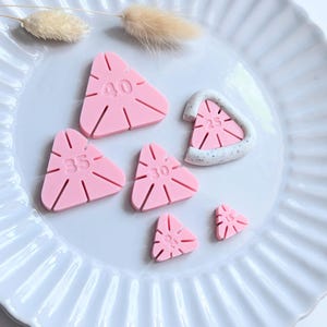 Könnte beinhalten: Eine Ansammlung von rosa, dreieckigen Objekten mit aufgedruckten Zahlen, angeordnet auf einem weißen, geriffelten Teller. Die Zahlen sind 15, 25, 30, 35 und 40. Einige haben einen weißen Rand. Trockenblumen sind im Hintergrund.