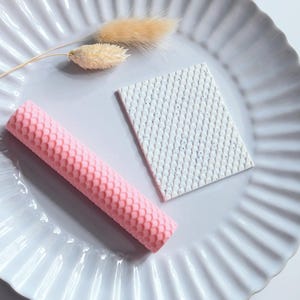 Peut inclure: Un objet cylindrique rose texturé et un objet rectangulaire blanc texturé sont présentés sur une assiette blanche cannelée. Des herbes décoratives beiges séchées agrémentent la composition. Les objets ont un motif en nid d'abeille.