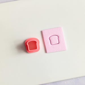 May include: A pink bread-shaped cookie cutter and a pink bread-shaped stamp are displayed on a white surface. The cookie cutter is a darker shade of pink, while the stamp is a lighter shade. The items are likely used for baking or crafting.