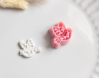 Puede incluir: Dos cortadores de galletas con forma de paloma, uno blanco y otro rosa, están sobre un plato blanco. La paloma blanca tiene un diseño de alas detallado. El cortador rosa tiene un contorno sólido y un interior detallado. También hay flores beige secas en el plato.