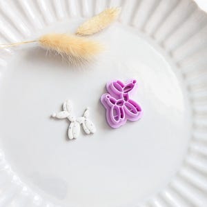 May include: A white balloon dog-shaped cookie cutter next to a white balloon dog-shaped cookie. The cookie cutter is purple. The cookie has small grey speckles. A sprig of dried, fluffy beige grass is also in the image.