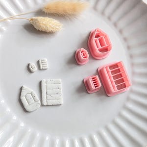 May include: A selection of pink polymer clay cutters in shapes like a rose, rectangle, and pencil, displayed on a white plate with a decorative rim. Small white clay shapes are also visible. A dried, beige plant stem is placed nearby.