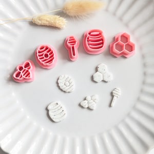 Puede incluir: Un juego de cinco cortadores de galletas de plástico rosa con formas de abeja, colmena, cuchara para miel, tarro de miel y panal, acompañados de cinco figuras de arcilla blanca con motas cortadas con estas herramientas.