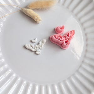 May include: A white ceramic plate with a fluted rim holds a small, white owl-shaped object, a pink butterfly-shaped object, and a small, pink, circular object. A sprig of dried, beige-coloured grass sits on the plate.