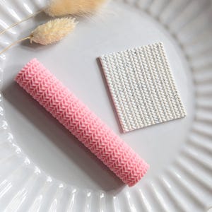 May include: A pink, textured, cylindrical object and a white, rectangular object with a knitted pattern are displayed on a white, fluted plate. Two dried, beige-coloured flower stems are also present, adding a touch of natural decor.