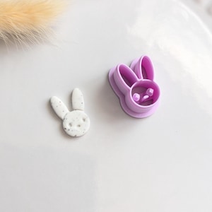 Peut inclure: Un emporte-pièce en forme de lapin violet clair à côté d'une petite figurine en argile en forme de lapin moucheté. L'emporte-pièce est en plastique et a un visage détaillé. La figurine en argile est blanche avec des mouchetures grises.