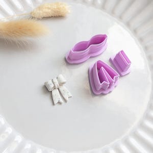 Könnte beinhalten: Ein weißer Keramikteller mit einer kleinen, verzierten Schleife und drei hellvioletten Ausstechern. Die Schleife ist weiß mit kleinen blauen Sprenkeln. Die Ausstecher haben die Form einer Schleife, eines gefalteten Bandes und eines Rechtecks.
