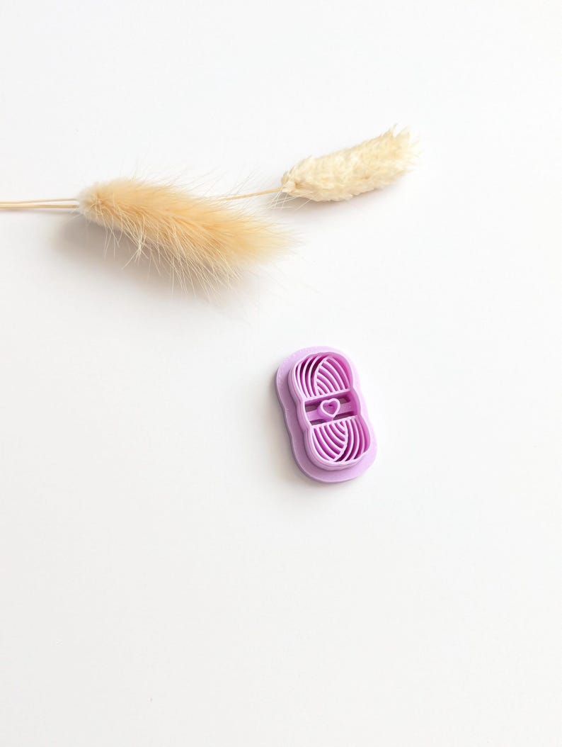 Puede incluir: Un peque&ntilde;o cortador de galletas en forma de bola de hilo de color lavanda con un coraz&oacute;n en el centro. El cortador es de pl&aacute;stico y tiene forma rectangular. Una ramita de hierba seca de color beige est&aacute; en el fondo.