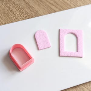 Könnte beinhalten: Drei rosa bogenförmige Objekte auf einer weißen Oberfläche. Eines ist ein Kunststoffschneider, die anderen beiden sind ausgeschnittene Formen. Die Formen haben eine strukturierte Oberfläche. Die Objekte sind auf einer weißen Oberfläche mit Holzgrund angeordnet.