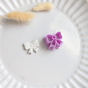 May include: A light purple plastic bow-shaped cookie cutter alongside a small, white, speckled bow-shaped clay piece. The cutter features a hollow centre with defined edges for shaping, while the clay piece displays a smooth, matte finish.