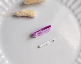 Puede incluir: Un plato blanco con borde acanalado contiene un pequeño cortador de galletas de color púrpura claro y un pequeño objeto blanco en forma de cuchillo con motas grises. Dos tallos beige secos y esponjosos también están en el plato.