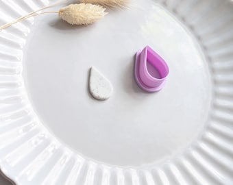 Könnte beinhalten: Ein weißer Keramikteller mit geriffeltem Rand enthält ein kleines, gesprenkeltes, tropfenförmiges Objekt, einen lila tropfenförmigen Ausstecher und zwei getrocknete, beige Blütenstiele. Die Objekte sind auf dem Teller vor weißem Hintergrund angeordnet.