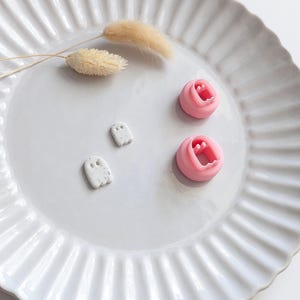 Op de afbeelding: Twee kleine witte kleistukjes in de vorm van een spookje met donkere spikkels en twee roze plastic koekjesvormen met een spookomtrek. De spookvormen hebben twee ronde uitsparingen voor de ogen en een geschulpte onderrand.