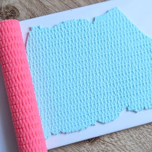 Puede incluir: Un rodillo rosa texturizado junto a una lámina de arcilla azul claro con un patrón tejido. La arcilla está sobre una superficie blanca, con un fondo de madera.