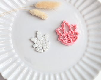 May include: A pink plastic cookie cutter shaped like two bells tied with a bow and surrounded by holly leaves, alongside a white speckled clay cutout of the same bell and holly design, both resting on a white fluted plate with two dried grasses.