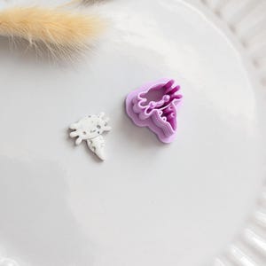 May include: Two cookie cutters, one carrot-shaped and the other flower-shaped, are on a white plate. The carrot cutter is white with grey spots. The flower cutter is purple. A dried, beige plant stem is in the background.