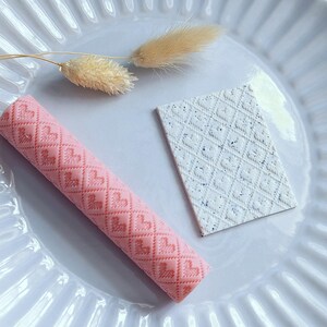 Puede incluir: Un rodillo rosa y un cuadrado blanco texturizado, ambos con patrones de corazones, descansan sobre un plato blanco acanalado. Un tallo de planta beige seco también está en el plato. El rodillo es cilíndrico.