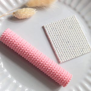 Op de afbeelding: Een roze, getextureerd, cilindrisch object en een wit, rechthoekig object met een gespikkeld patroon worden gepresenteerd op een wit, gegroefd bord. Twee gedroogde, beige bloemstengels zijn ook aanwezig en voegen een vleugje natuurlijk detail toe aan de compositie.