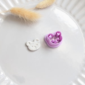 May include: A white, bear-shaped clay earring next to a purple bear-shaped cookie cutter with heart details. The earring is on a white plate with a dried plant stem.