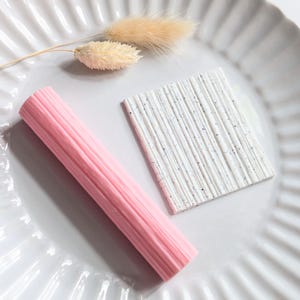 May include: A pink, ribbed, cylindrical object and a white, speckled, rectangular object are displayed on a white, fluted plate. A dried, beige plant stem is also on the plate. The objects appear to be decorative or artistic.