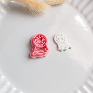 May include: A pink cat-shaped cookie cutter and a white cat-shaped fondant cutter are on a white plate. The pink cutter has a detailed cat design. The white cutter has a simplified cat design. Dried beige decorative grass is in the background.