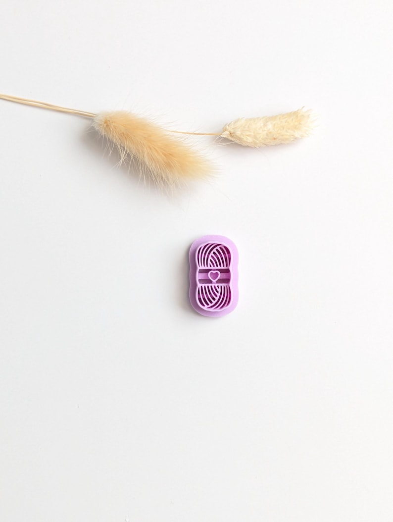 Puede incluir: Cortador de galletas morado en forma de ovillo de lana con un coraz&oacute;n en el centro. El cortador est&aacute; sobre una superficie blanca con un tallo de planta beige seco y cabezas de semillas esponjosas.