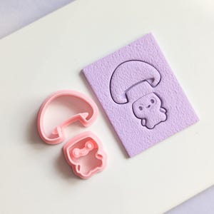 Könnte beinhalten: Zwei rosafarbene Plastik-Ausstecher in Pilzform. Ein Ausstecher ist ein einfacher Pilz, der andere ist ein Cartoon-Pilz mit einem Gesicht. Die Ausstecher befinden sich auf einer weißen Oberfläche.