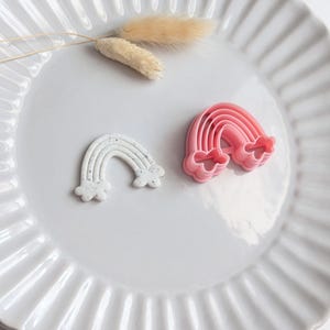 May include: Two rainbow-shaped cookie cutters, one white with speckled details and the other pink, are displayed on a white plate. A sprig of dried, beige-coloured flowers is also on the plate. The plate has a ridged design.