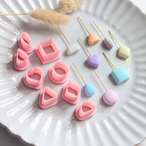 May include: A collection of pink plastic geometric clay cutters in heart, square, circle, and teardrop shapes alongside several small, pastel-coloured clay beads attached to thin gold metal pins, including speckled, marbled, and solid matte finishes.