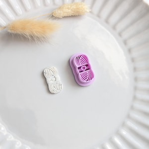 Pode incluir: Dois cortadores de biscoitos em forma de fio, um branco e outro lavanda, estão em um prato branco. O cortador lavanda tem uma forma de coração no centro. Um raminho de grama bege seca também está no prato.