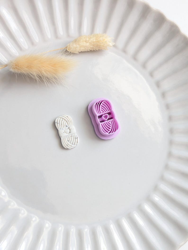 Puede incluir: Dos cortadores de galletas con forma de hilo, uno blanco y otro lavanda, est&aacute;n sobre un plato blanco. El cortador lavanda tiene forma de coraz&oacute;n en el centro. Una ramita de hierba beige seca tambi&eacute;n est&aacute; en el plato.