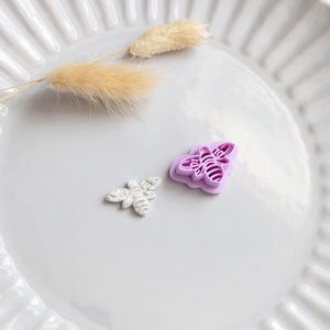 May include: A white ceramic plate with a fluted edge holds a small, white bee-shaped object and a purple bee-shaped cookie cutter. Dried, fluffy beige grass stems are also on the plate.
