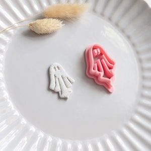 May include: A white ceramic plate displays a small, speckled white ghost and a pink ghost-shaped cookie cutter. A dried, beige plant stem is also on the plate. These items are likely for Halloween baking or crafts.