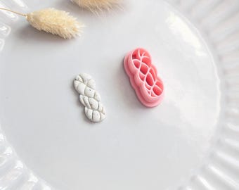 Puede incluir: Un plato de cerámica blanca con borde acanalado contiene una pequeña pieza de arcilla trenzada gris claro y un cortador de arcilla trenzada rosa. Dos tallos de flores beige secas y esponjosas también están en el plato.