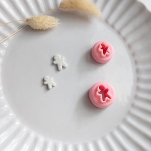 May include: Two pink circular clay cutters with a gingerbread person shape in the centre, accompanied by two small white speckled gingerbread person clay cutouts. The items are arranged on a white fluted plate with two dried beige grass stems.