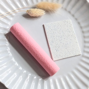 May include: A pink, cylindrical item and a white, rectangular item with blue speckles are presented on a white, fluted plate. Dried, beige-coloured flowers are also present, creating a minimalist aesthetic.