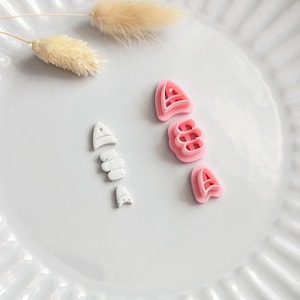May include: A white ceramic plate displays several small cookie cutters. Pink cutters are shaped like the letter "A" and a cluster of three rounded shapes. White cutters are in the shape of a fish. Dried flowers are also present.