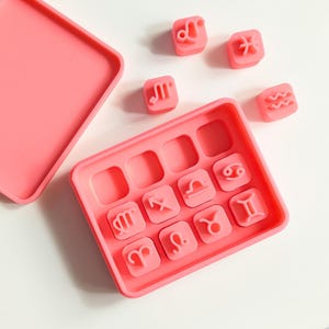 May include: A pink silicone mould with 12 square cavities, each imprinted with a different zodiac symbol. Several loose zodiac symbol stamps are scattered around the mould. The mould has a matching lid.