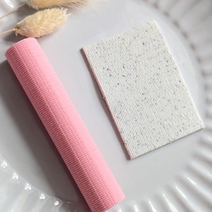 Könnte beinhalten: Ein rosa, geripptes, zylindrisches Objekt und ein rechteckiges, gesprenkeltes, weißes Objekt werden auf einem weißen, geriffelten Teller präsentiert. Getrocknete, beige-farbene Blumen sind ebenfalls vorhanden und verleihen eine natürliche Dekoration.