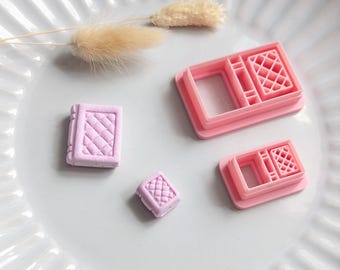 May include: A collection of pink cookie cutters and a small, lavender-colored cookie on a white plate. The cutters are in the shape of a book and a window. The cookie has a quilted pattern.