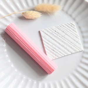 May include: A pink, ribbed, cylindrical object and a white, square object with diagonal lines and speckled details are arranged on a white, fluted plate. A sprig of dried, beige flowers is also present. The objects are likely decorative or edible.