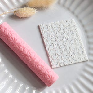May include: A pink, cylindrical item and a white rectangular item with a floral pattern are displayed on a white, ridged plate. A dried, beige plant stem is also present, adding a natural element to the composition.