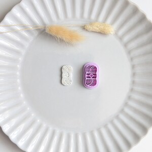 Puede incluir: Dos cortadores de galletas con forma de hilo, uno blanco y otro morado, est&aacute;n sobre un plato blanco con volantes. El cortador morado tiene un dise&ntilde;o de coraz&oacute;n. Tambi&eacute;n hay tallos beige secos y esponjosos en el plato.