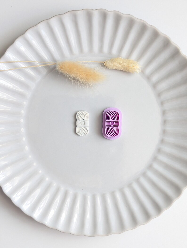 Puede incluir: Dos cortadores de galletas con forma de hilo, uno blanco y otro morado, est&aacute;n sobre un plato blanco con volantes. El cortador morado tiene un dise&ntilde;o de coraz&oacute;n. Tambi&eacute;n hay tallos beige secos y esponjosos en el plato.